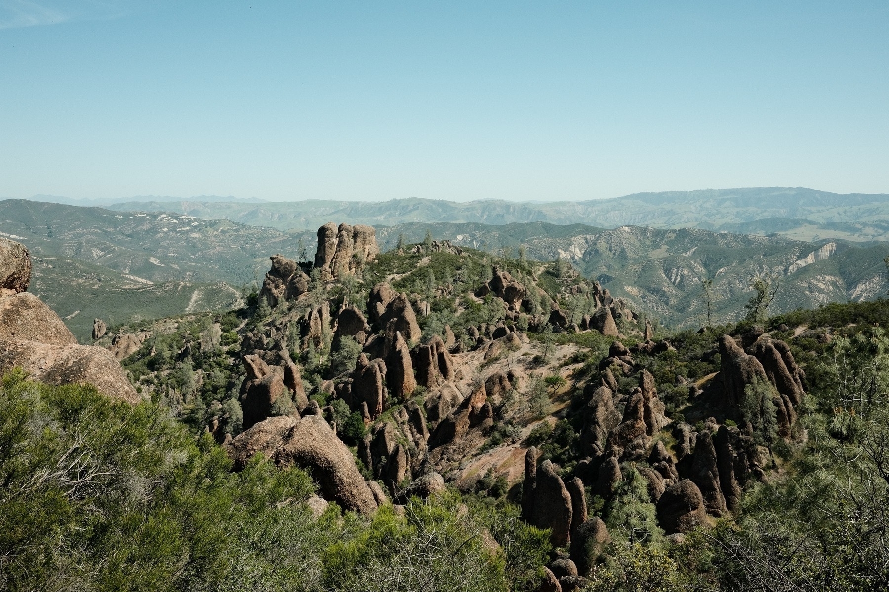 Pinnacles of rock jut out of a hilly landscape, with their northsides in shadow and southern sides completely lit up. Vegetation is quite green with a classic kodachrome blue sky and mountain ranges along the horizon.