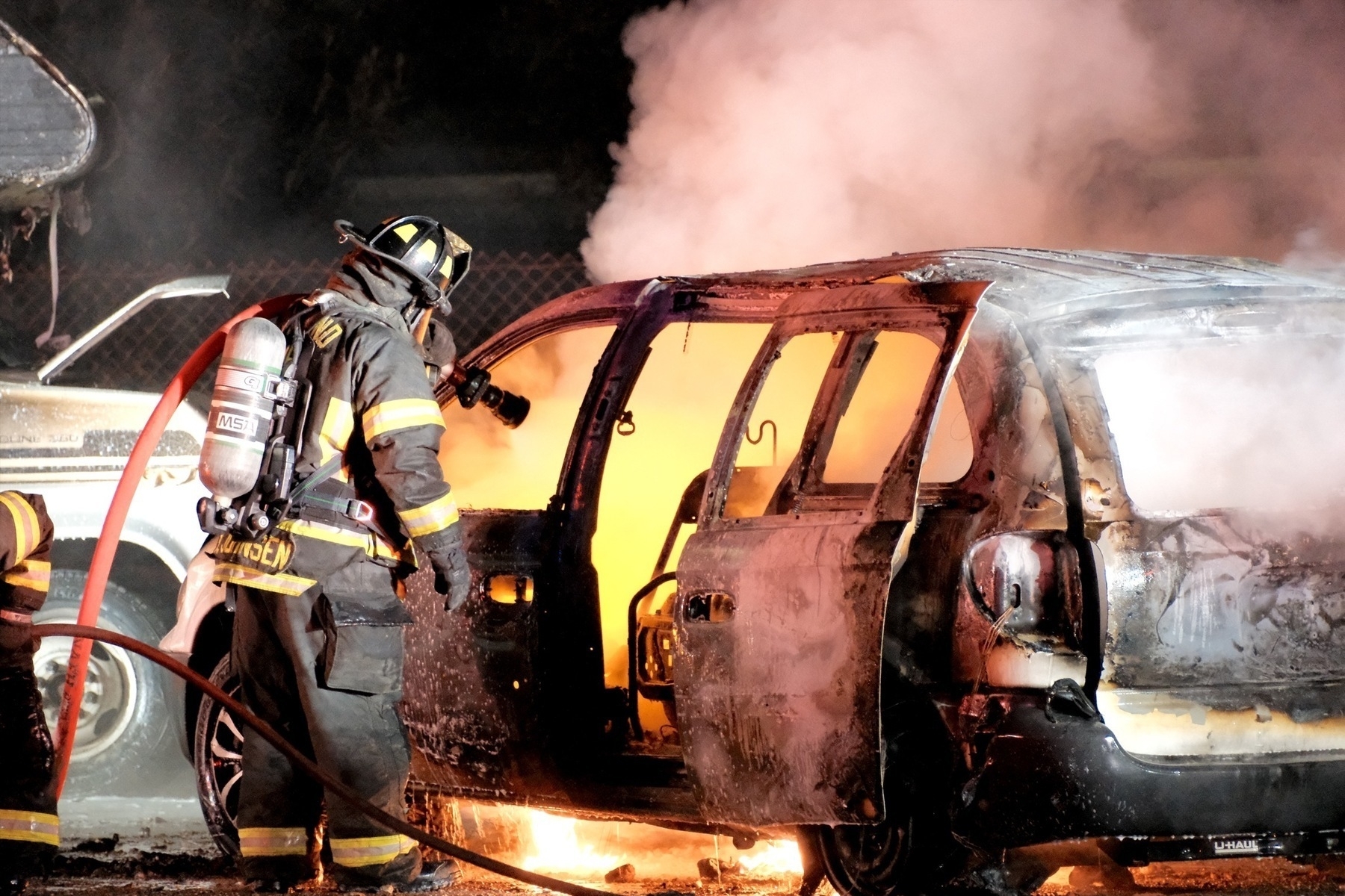 Fire fighter in full respirator equipment with a hose flooding the interior of the car that's on fire. Burning fluids drop below the car, like lava. The car is extremely charred.
