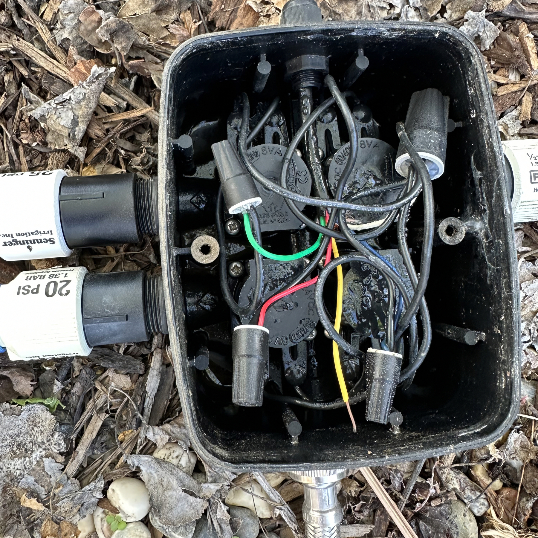A irrigation valve box with wires tied together by wire nuts including the green wire. The small box sits atop some gravel and has a stainless steel braided hose providing input water. There are 20-25 GPH pressure regulators and adapters to drip irrigation hoses.