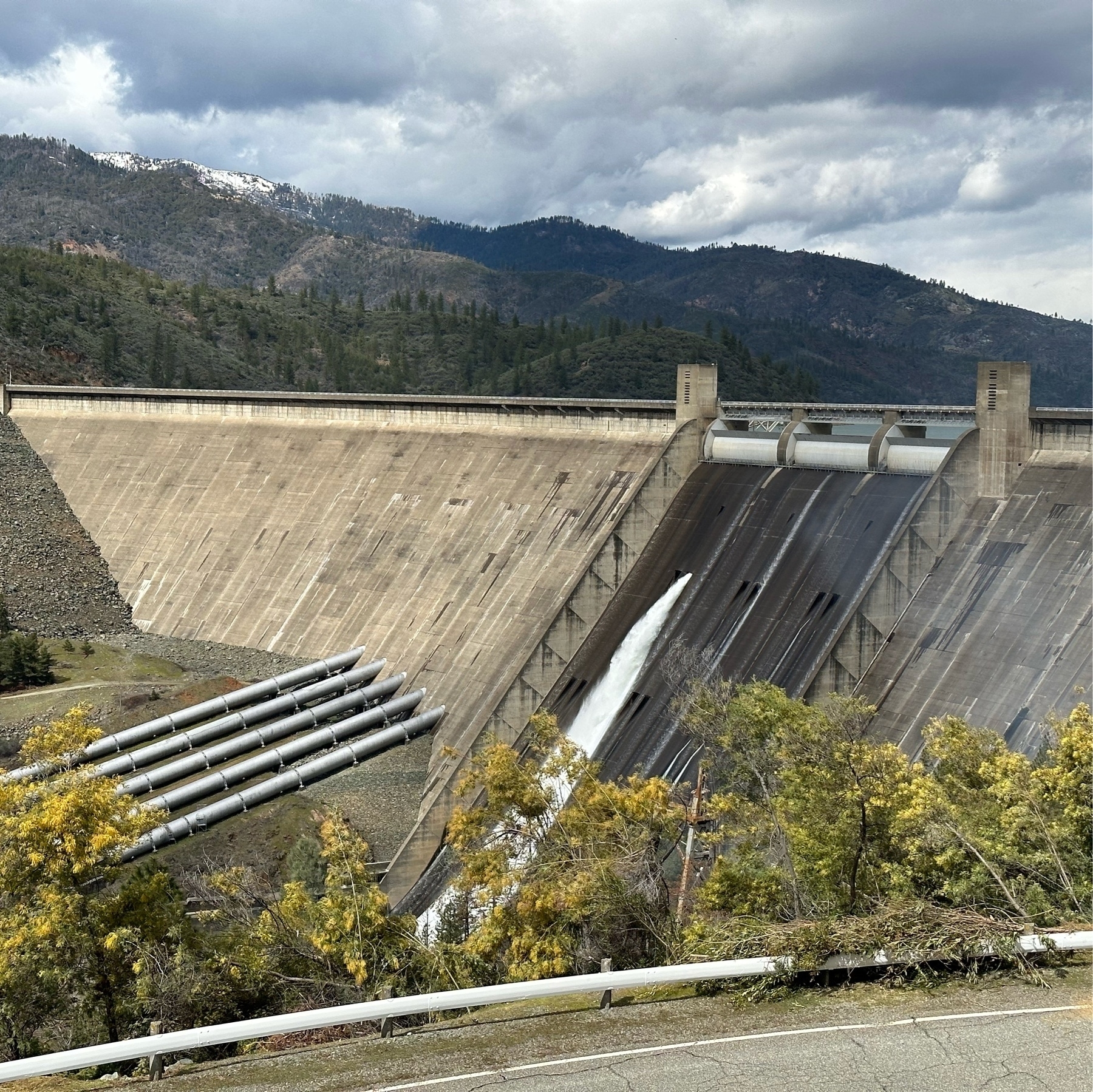 The massive shasta dam with snow capped mountains in the background. A spillway gate appears to be open, sending an enormous amount of water into the Sacramento River. It is an overcast day.
