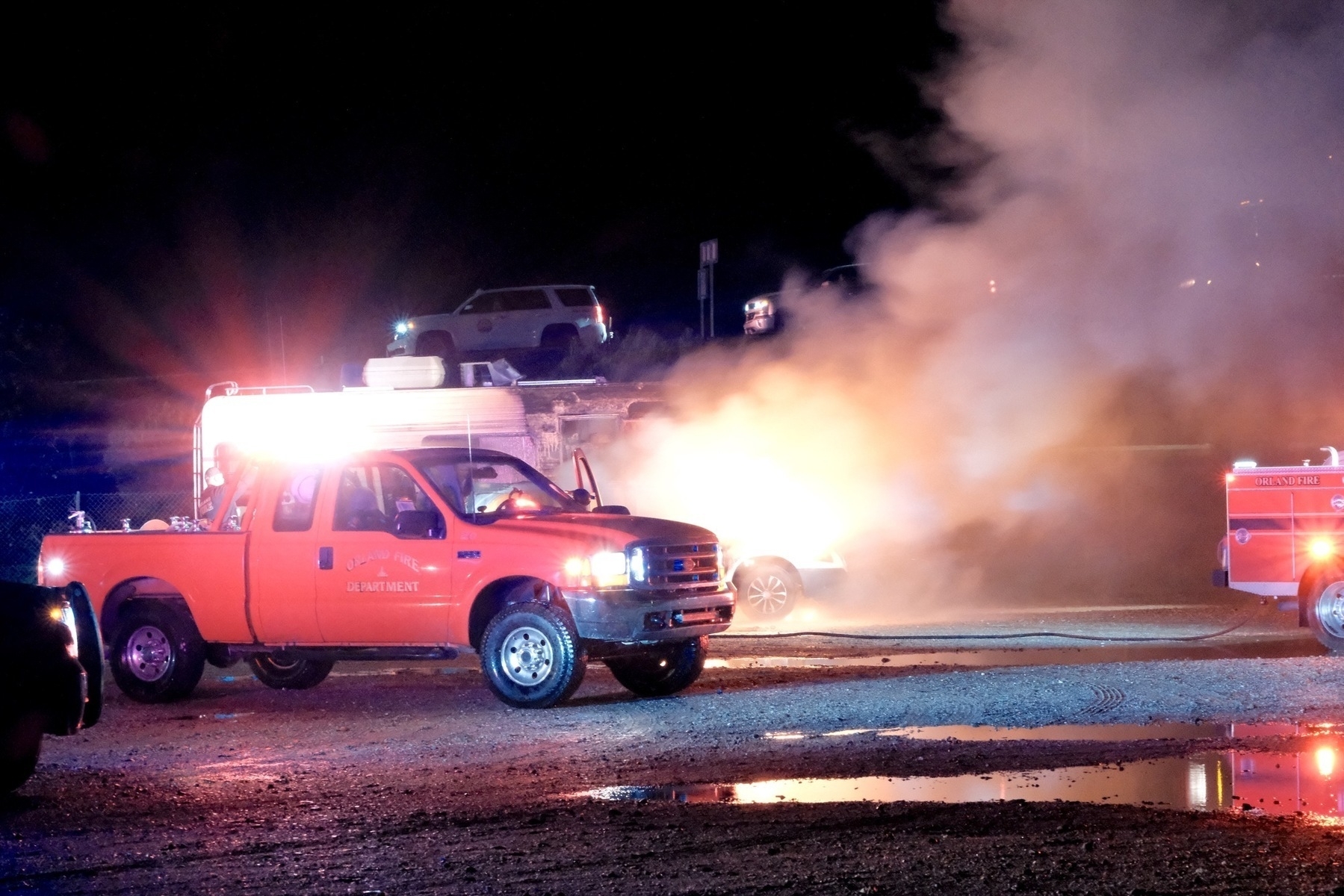 A wide angle view of the RV and car on fire, in front of an overpass. Two fire trucks are parked in front of the blaze on a gravel lot with some ponding.