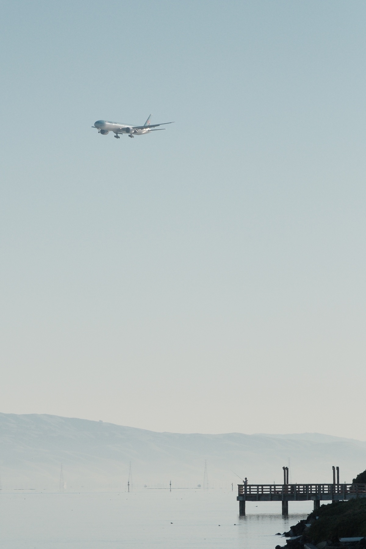 A mid-zoom of a Korean Air 777-3 HL7204 ICN-SFO on approach during a hazy day. There’s a silhouetted pier with somebody fishing at the very end if it. A hazy mountain is in the background. 