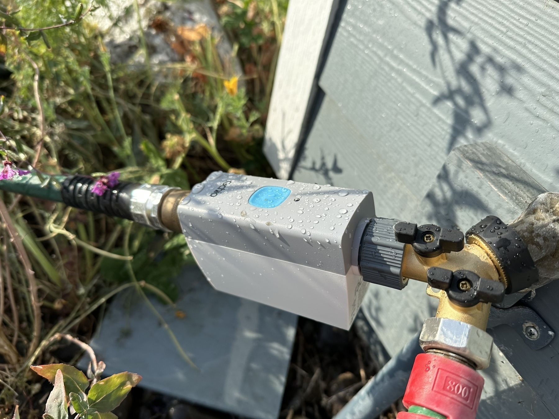 The grey and white Rachio hose sprinkler timer. It is outdoors and connected to a Y spigot. There’s a back flow preventer and a hose attached to it. It has some water on it.