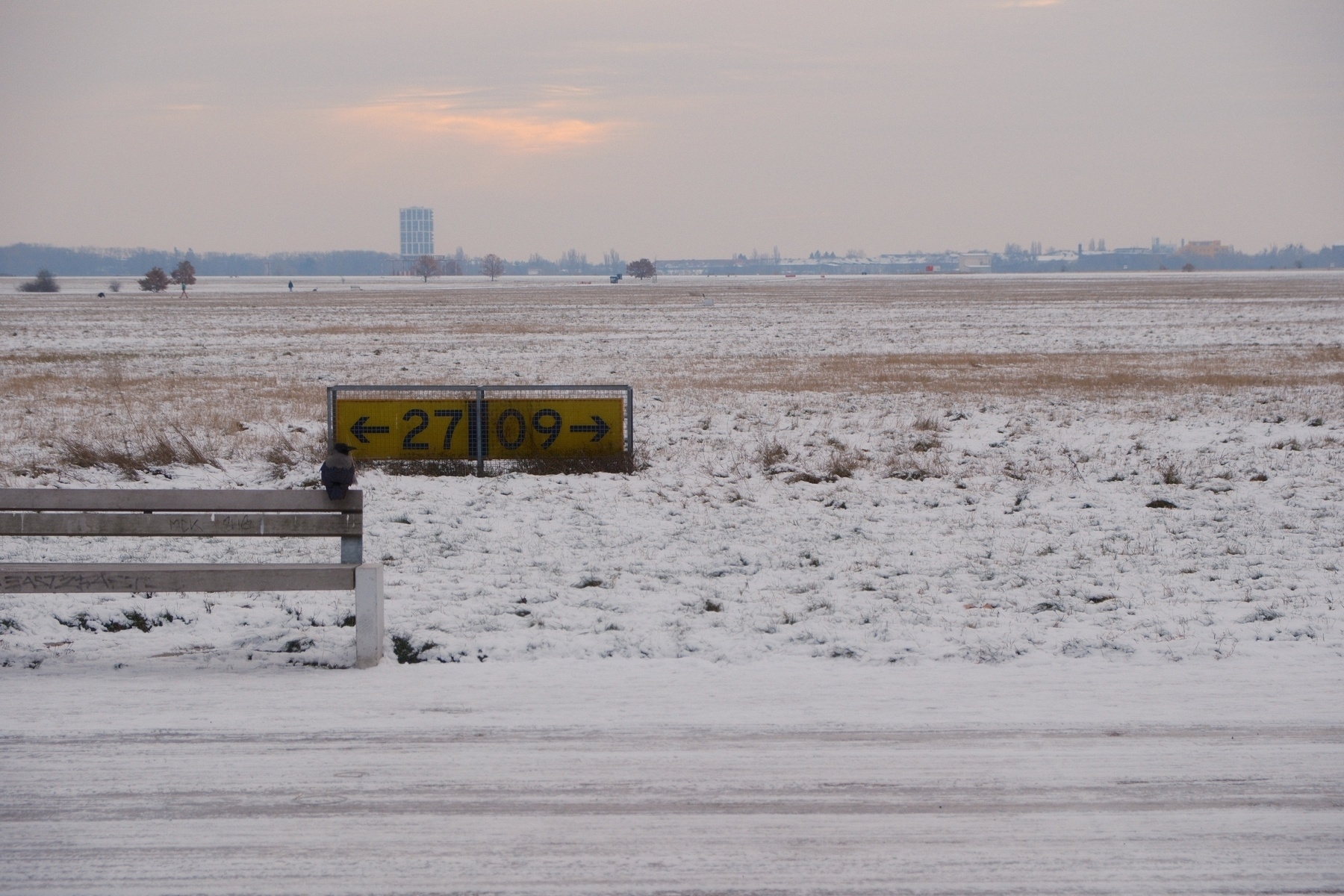 A snowy landscape with runway direction signs reading “27” with an arrow pointing left and “09” with an arrow pointing right, indicating runway headings at an airport. The background features a flat horizon with the sun trying to shine through an overcast sky and distant buildings. There’s a crow sitting on a bench in the foreground.