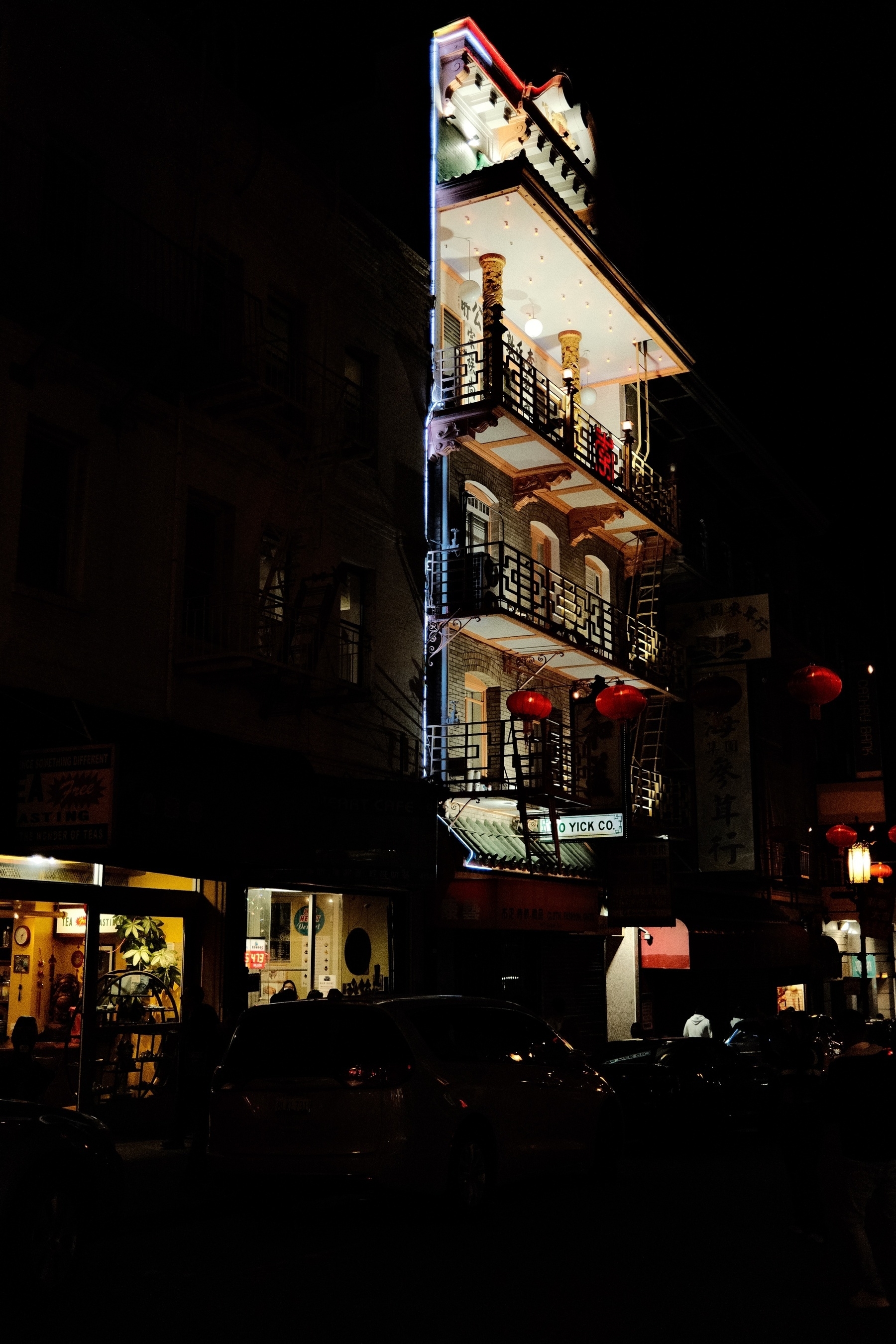 It is night in the city where there’s a five story building where the front is only lit up. Small li windows reveal shops are along the dark street. There are a few red unlit Chinese New Year lanterns strung across the street.