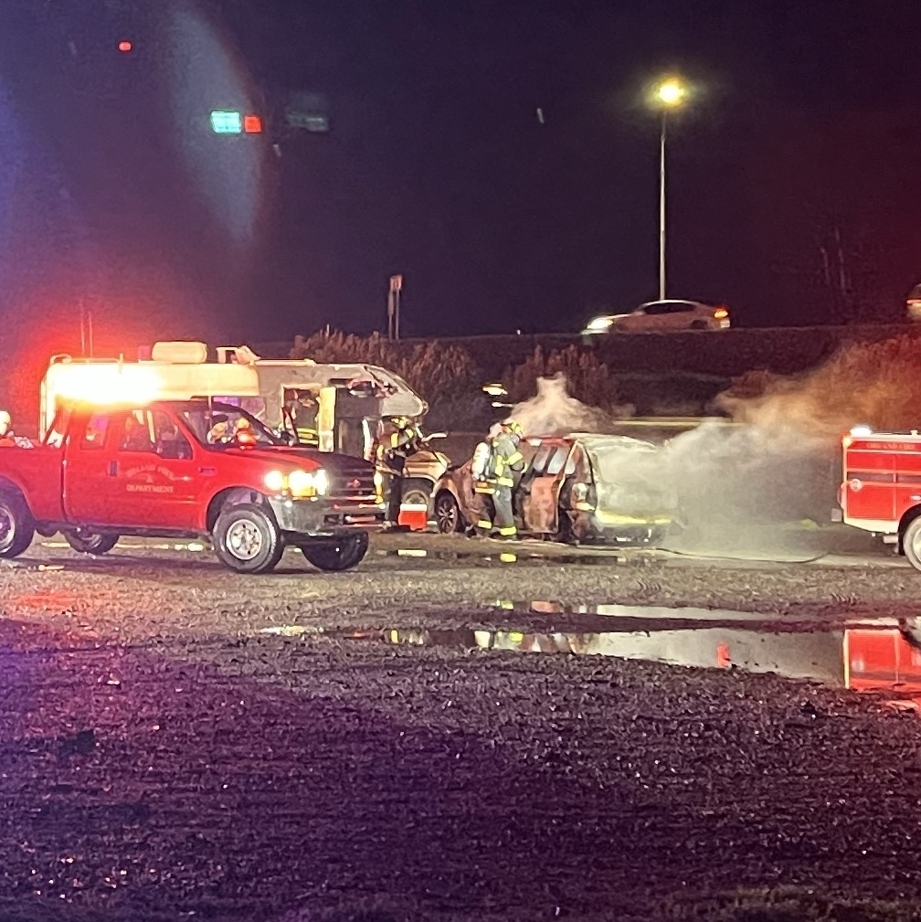 A night scene of fire trucks in front of a burnt out RV and car.