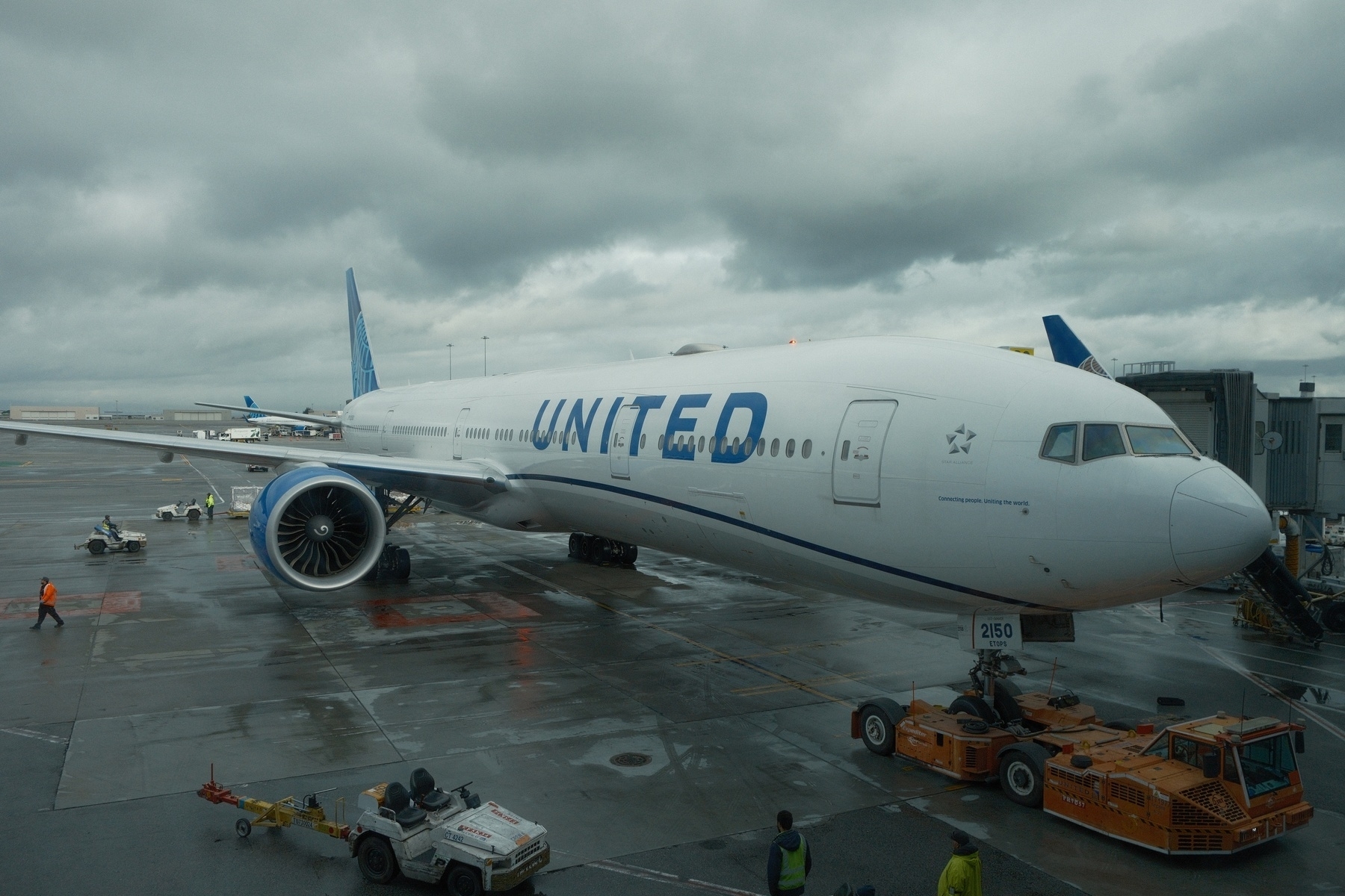 United 777-300 parked at SFO gate G9 just as it’s towed in. It’s a cloudy day and the tarmac is wet and shiny.