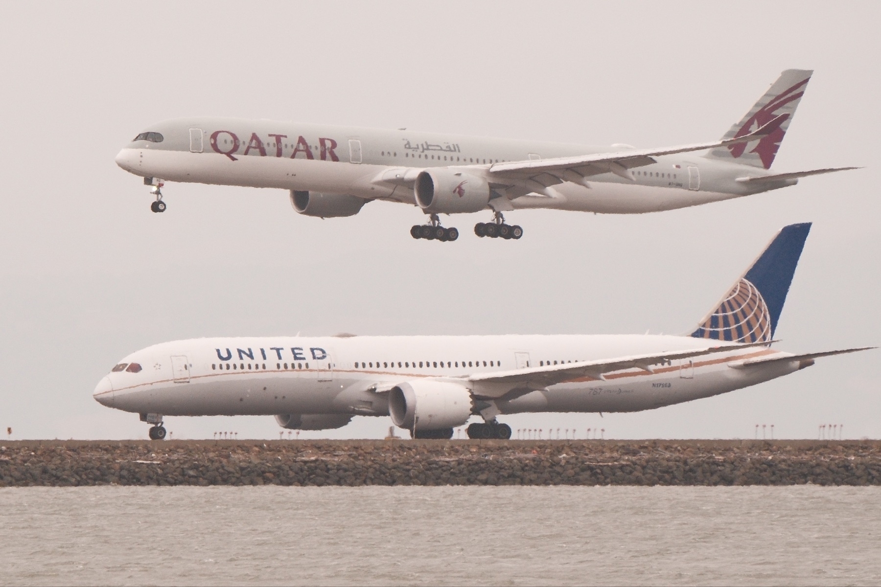 A United 787 prepares for take off, while above, a Qatar 777 airliner lands on an adjacent runway.