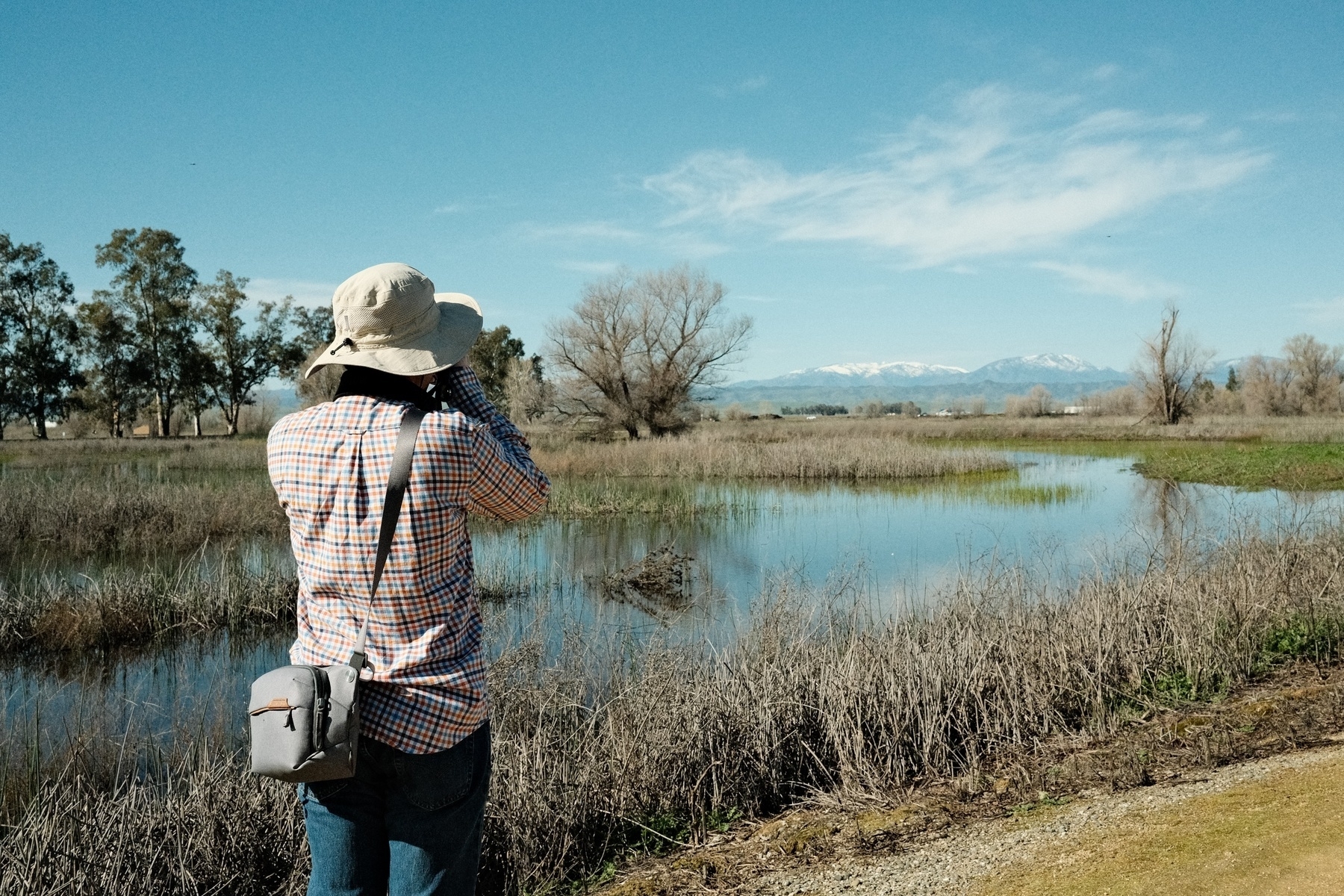 A person in a wide-brimmed hat and plaid shirt standing by a pond and looking through a camera at a landscape with distant snow-capped mountains and blue sky with wispy clouds. 