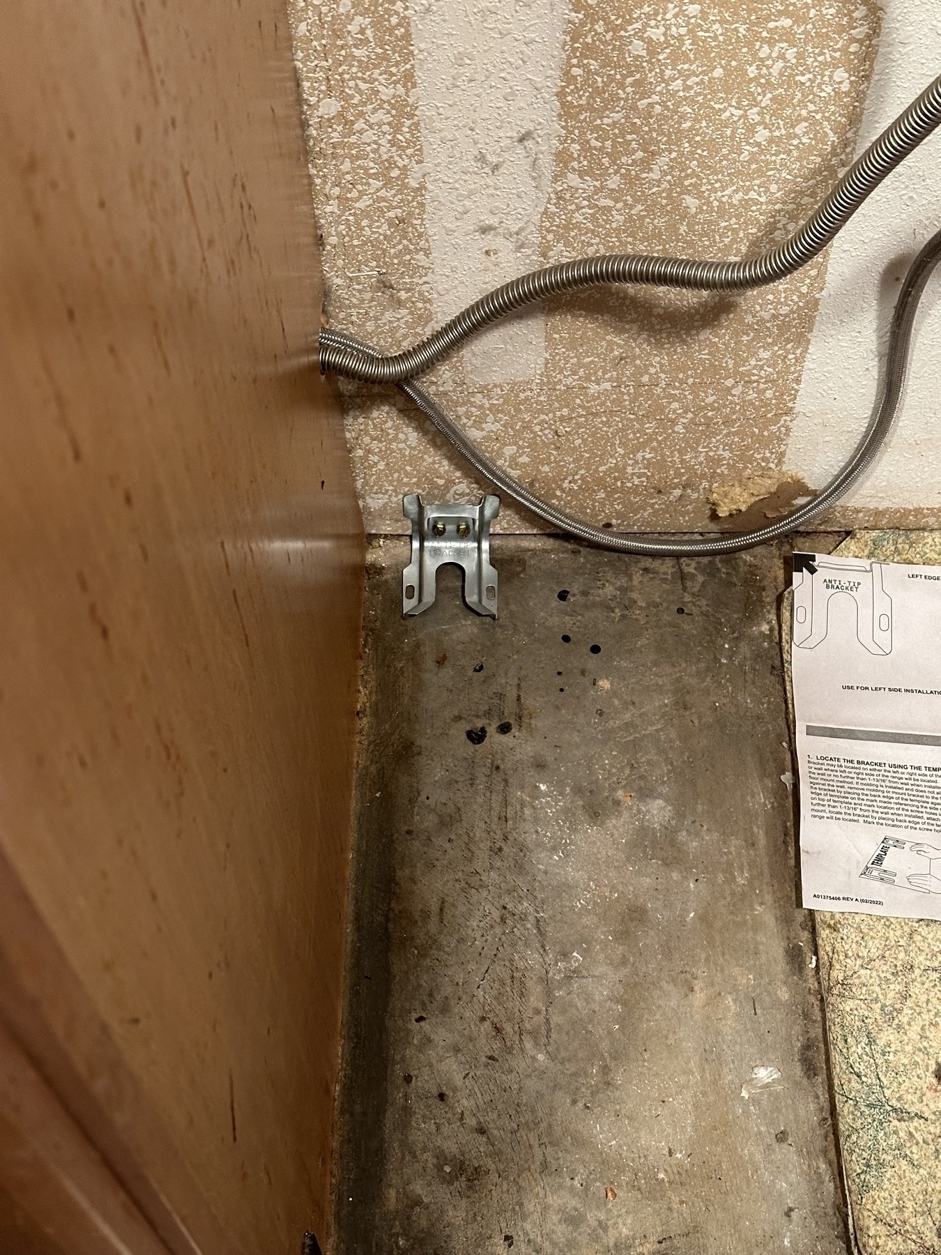 A corner behind a kitchen appliance with a wall and floor showing signs of dirt and residue. There are two metallic conduit pipe, a metal anti-tip bracket, and an installation instruction manual is partially visible on the right side.