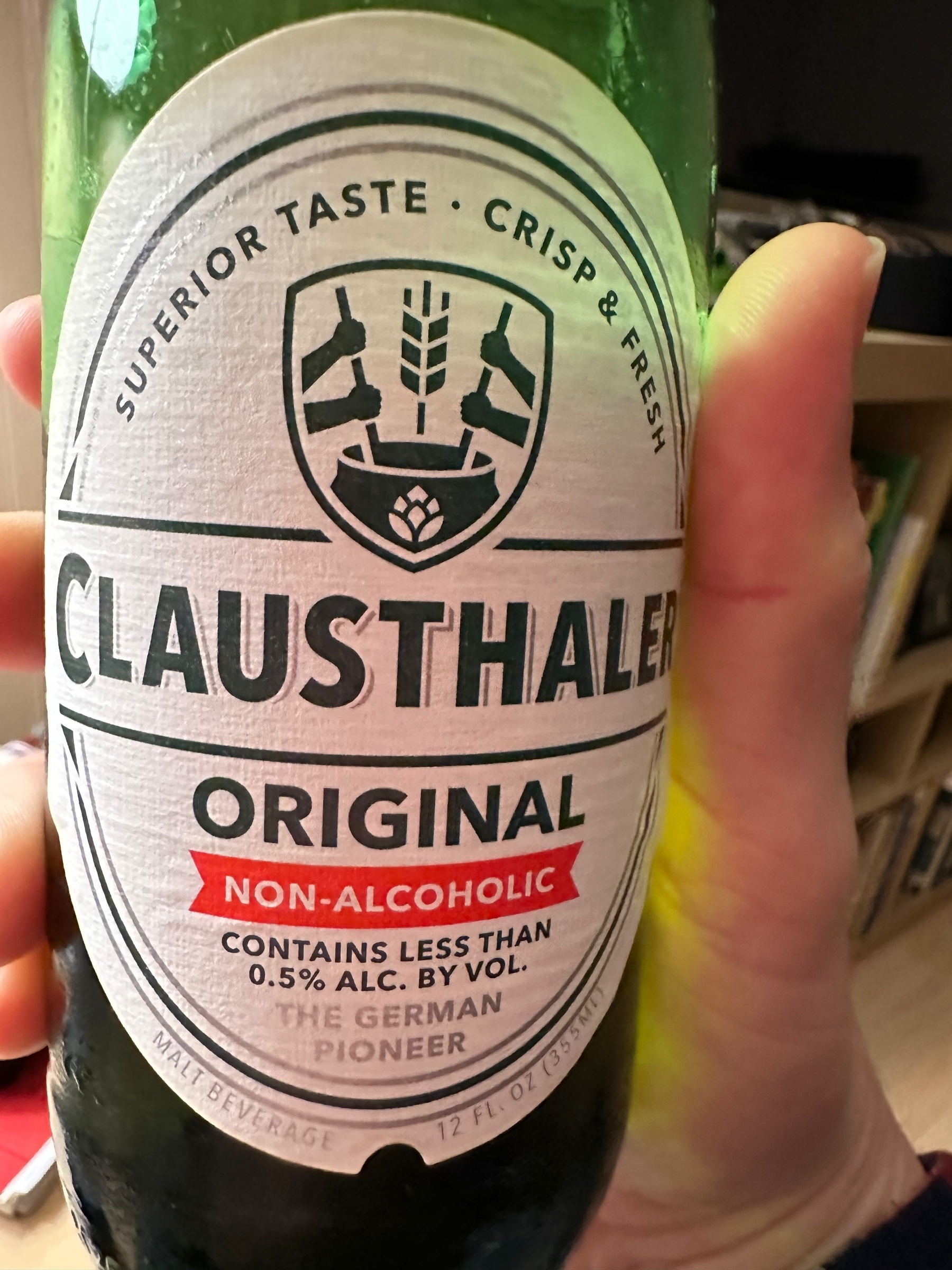 A hand holding a bottle of Clausthaler Original Non-Alcoholic beer, which contains less than 0.5% alcohol by volume. The label includes the phrases “Superior Taste,” “Crisp & Fresh,” and “The German Pioneer”