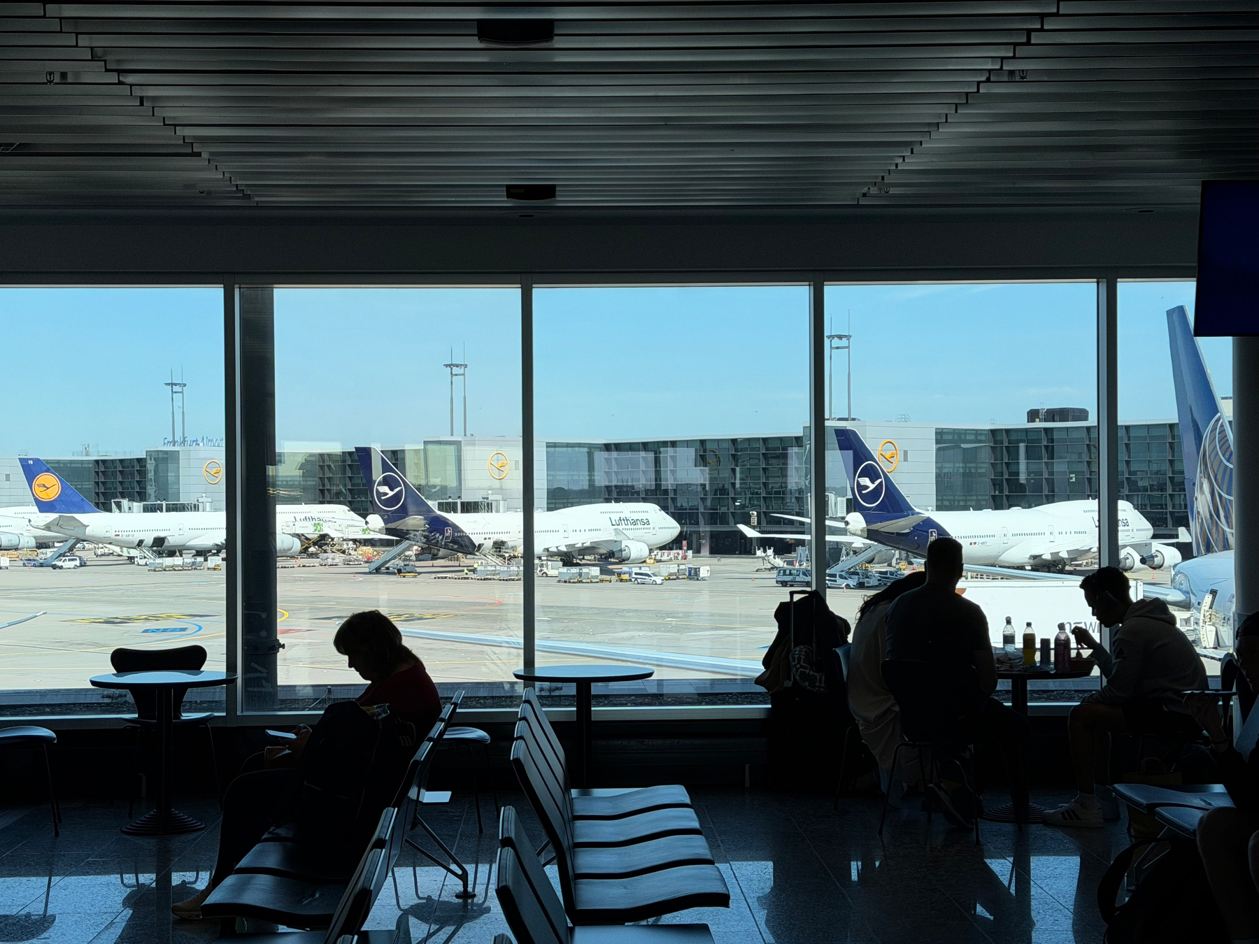 At an airport terminal, several silhouetted people sit facing windows with a view of threeLufthansa 747-8 aircraft on the tarmac.