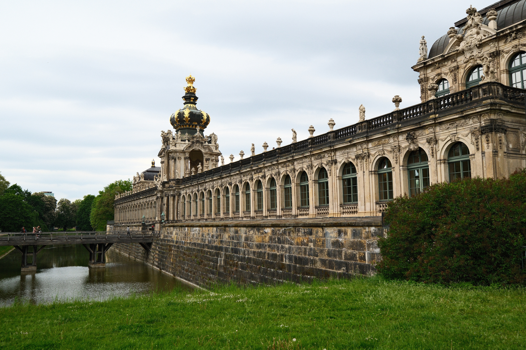 Auto-generated description: A grand, historic architectural structure with ornate details and a gold-crowned tower is seen alongside a grassy area and a waterway.