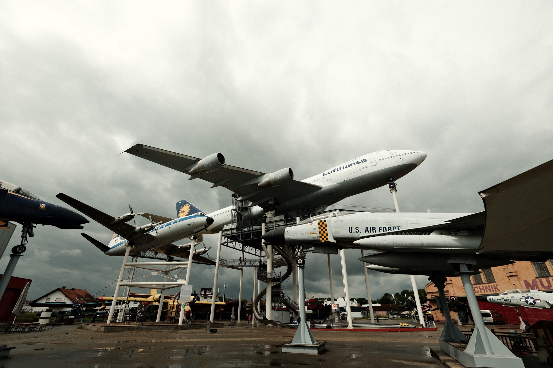 At least six different static display aircraft are in one shot. It's a cloudy day with slightly wet pavement. Most aircraft are mounted dozens of feet above on complicated metal scaffolds. 